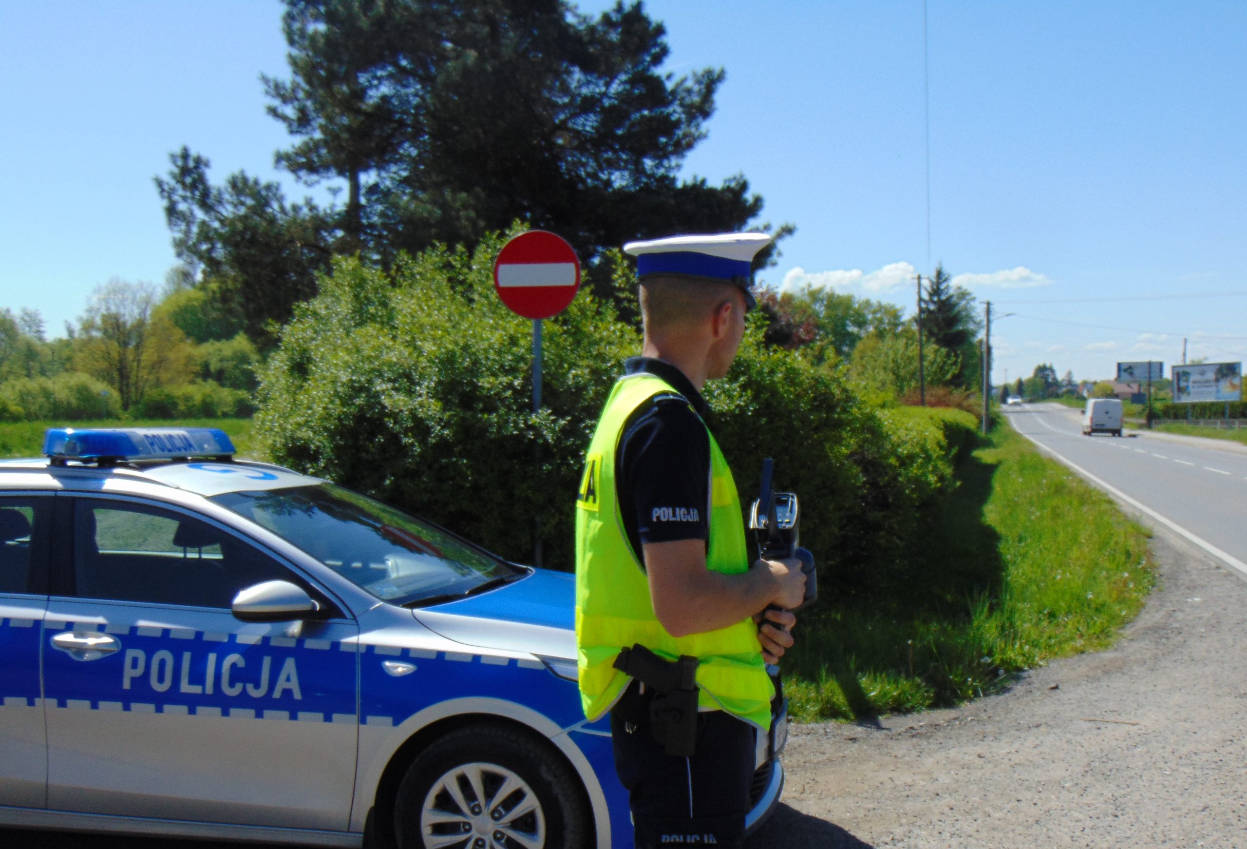 KPP Oświęcim policjant WRD czuwa nad bezpieczeństwem w ruchu drogowym. Obok policjanta stoi radiowóz Info Oswiecim